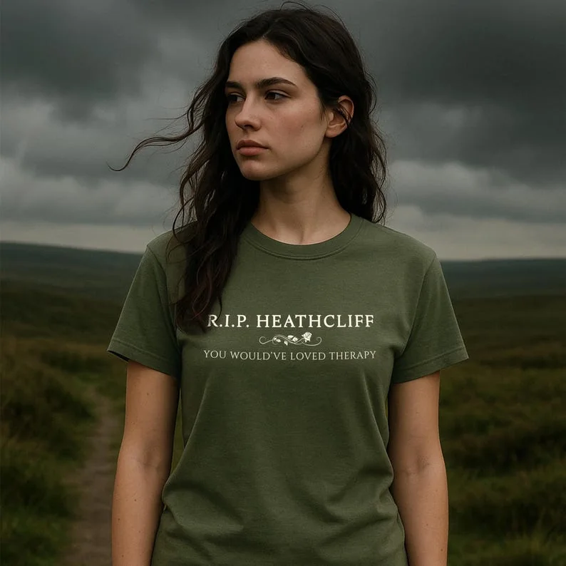 A feminine-presenting figure stands against a windswept hedge on a misty countryside path, wearing a Moss green Comfort Colors tee. The model’s wild hair and proud posture evoke Catherine Earnshaw. The landscape suggests Yorkshire moors, enhancing the gothic literary atmosphere.