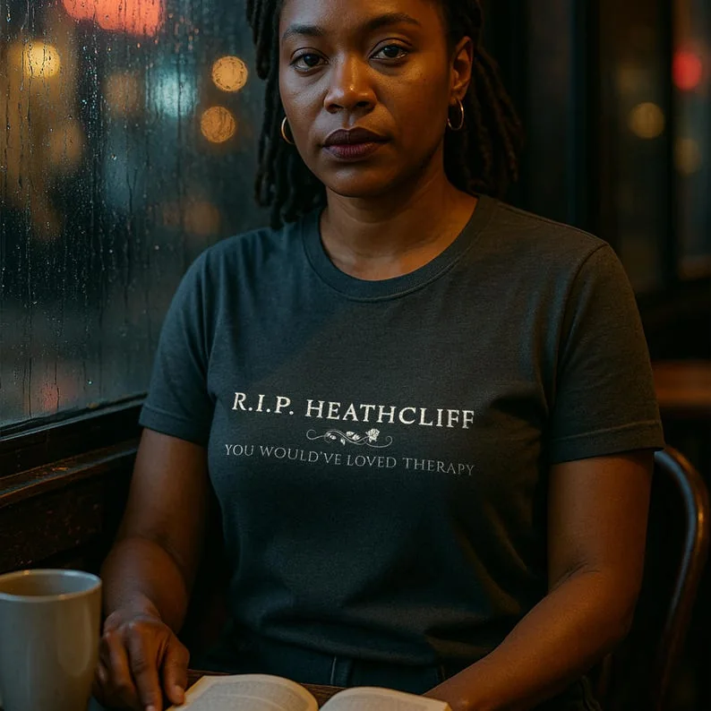A woman with dark, curly hair sits by a rain-streaked café window wearing a Pepper Comfort Colors t-shirt. Her expression is pensive, with a vintage novel open in front of her. The moody café lighting and raindrops on glass create a gothic, introspective vibe.