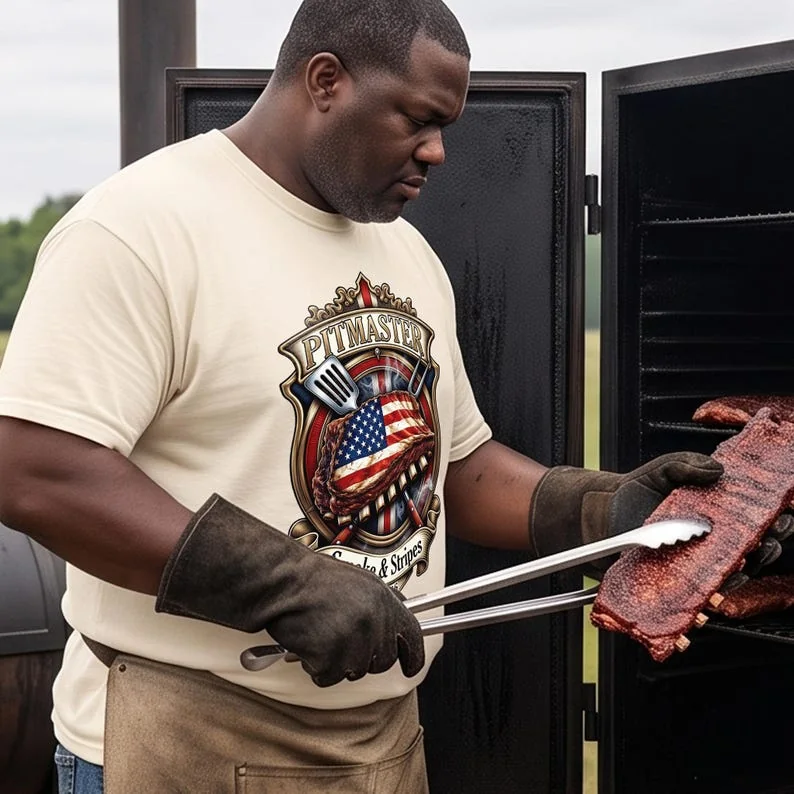 An outdoor lifestyle photo of a male pitmaster wearing a cream Ivory graphic shirt, using metal tongs to handle a rack of ribs on a large black commercial offset smoker pipe.