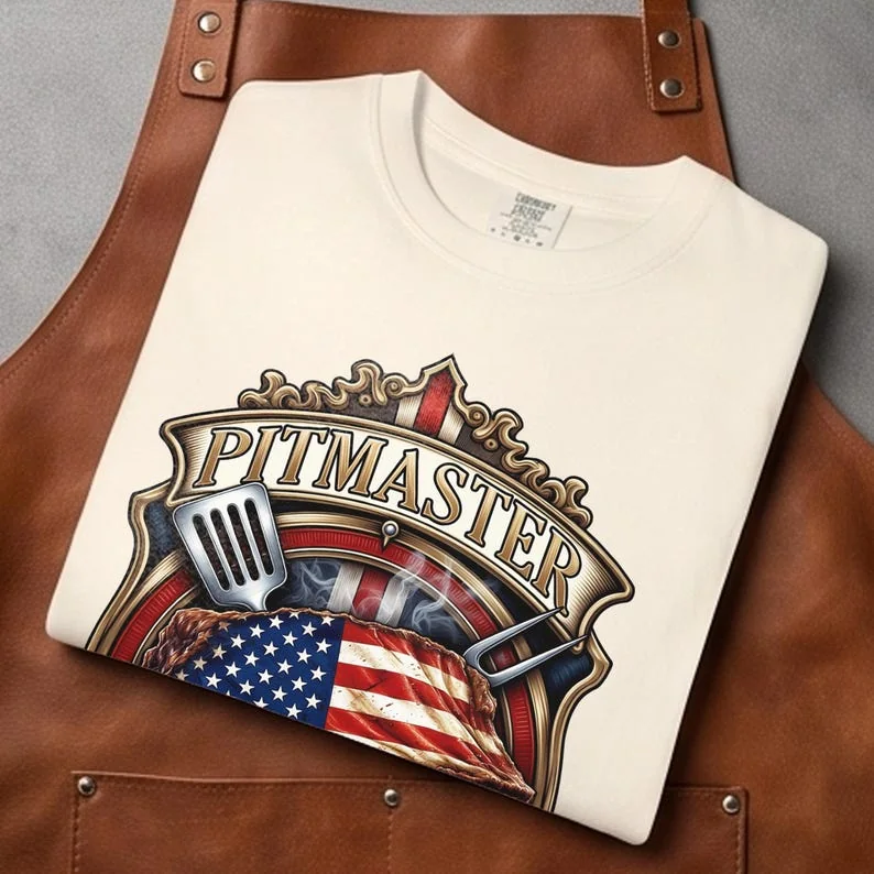 A close up flat lay of an Ivory off-white heavy cotton t-shirt folded neatly on top of a rugged brown leather pitboss apron, showcasing the institutional American heritage Pitmaster crest design.