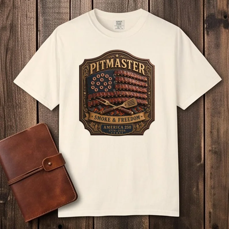 A cream-colored Ivory Comfort Colors 1717 t-shirt displayed flat on a dark wooden surface. The shirt features a vintage graphic design reading "PITMASTER SMOKE & FREEDOM" with an American flag composed of BBQ ribs. A brown leather journal sits next to the shirt.