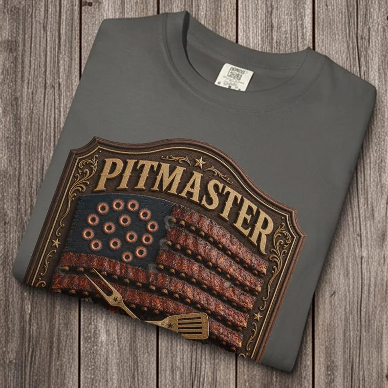 A charcoal gray Pepper Comfort Colors 1717 t-shirt folded neatly on a gray wooden plank background. The shirt features a detailed vintage graphic with an American flag made of BBQ ribs and the text "PITMASTER".